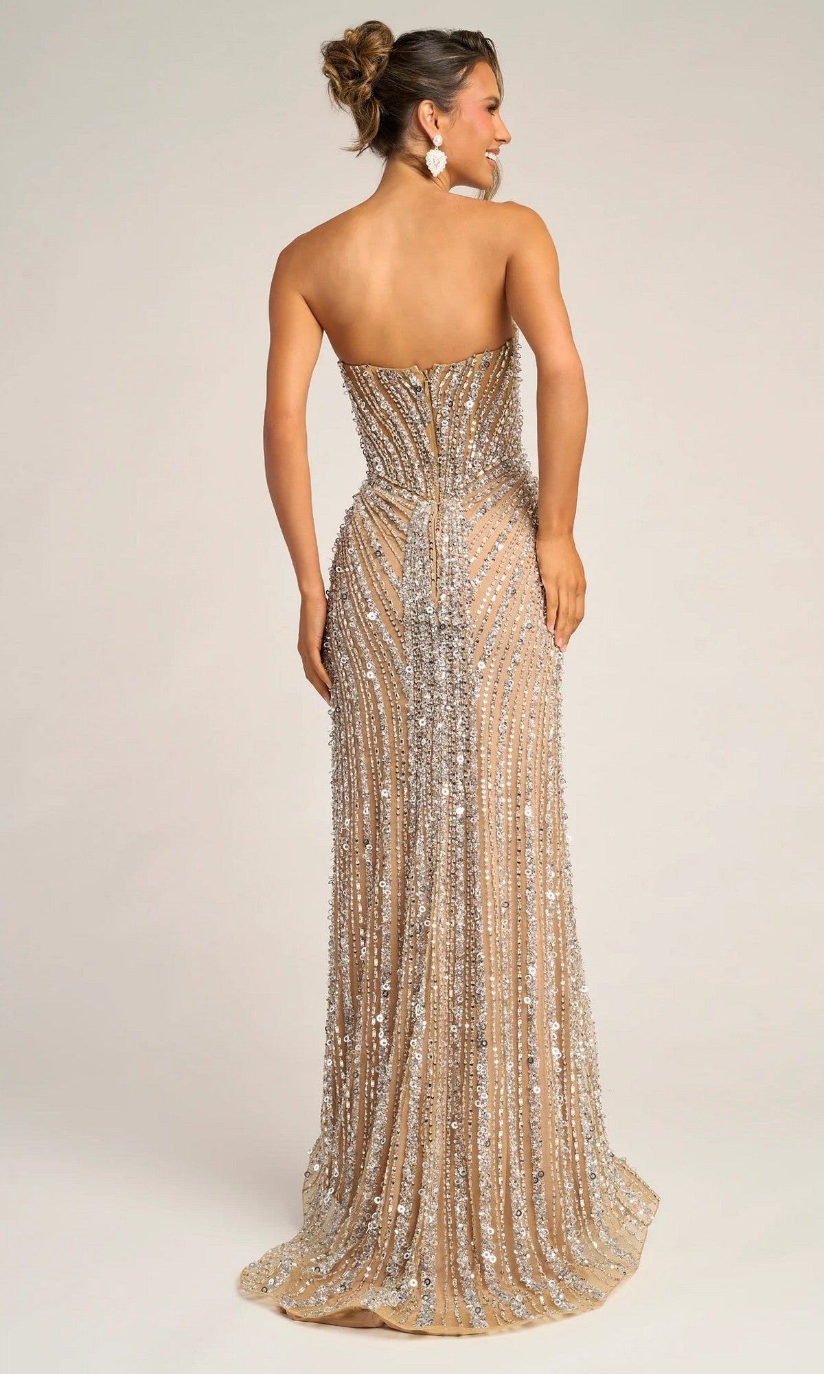 A woman with an elegant updo faces away, wearing the Portia and Scarlett PS26403C long formal dress featuring a strapless sweetheart neckline, floor-length nude fabric, and silver sequin embellishments.